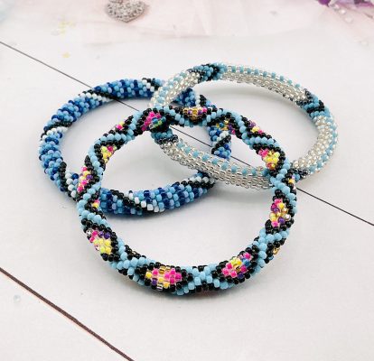 Three handmade seed bead bracelets in shades of blue, black, white, and vibrant neon colors, arranged on a light surface.